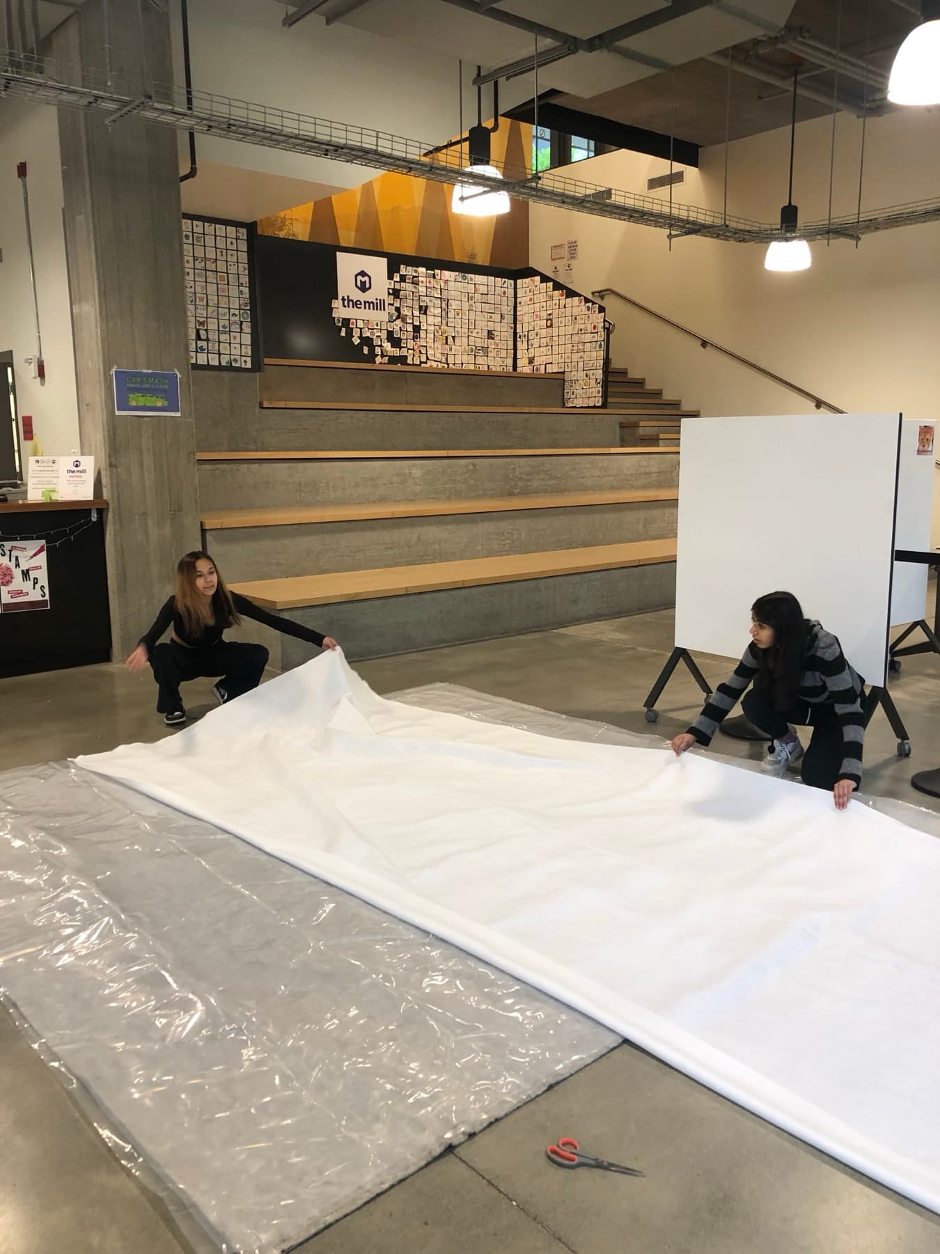 Two team members laying out the full white mat fabric over plastic sheeting on a makerspace floor.