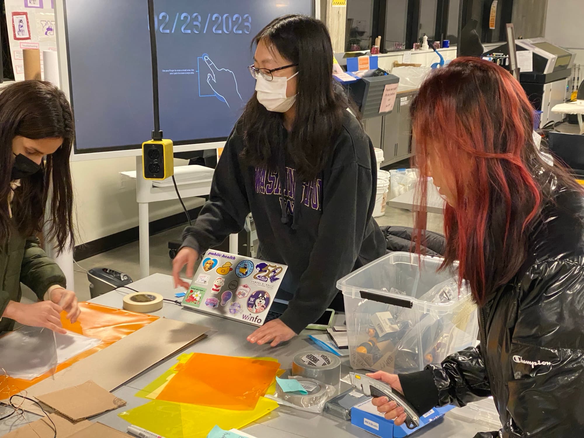 Three team members at a workbench testing coloured gel sheets, fabrics, and cardboard.