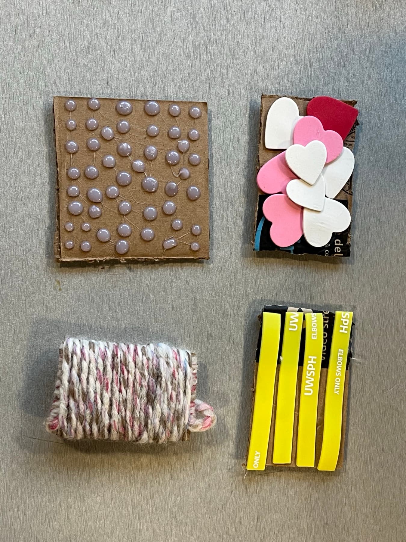 Four cardboard block stand-ins on a table, each wrapped in a different texture: rounded pins, foam hearts, wound yarn, plastic strips.